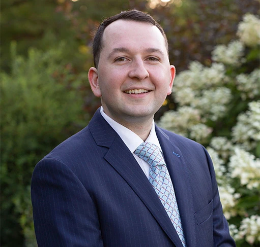 Doctor Kalloch professionally dressed in a dark suit and pale blue tie standing in a garden setting, looking at the camera with a warm smile.